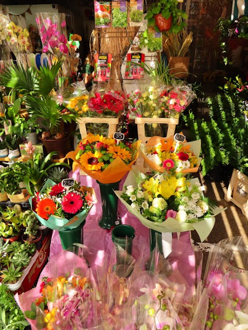 A vibrant display of fresh floral bouquets arranged on a pink cloth-covered table inside a flower shop, featuring a variety of flowers including bright yellow sunflowers, pink gerbera daisies, white c