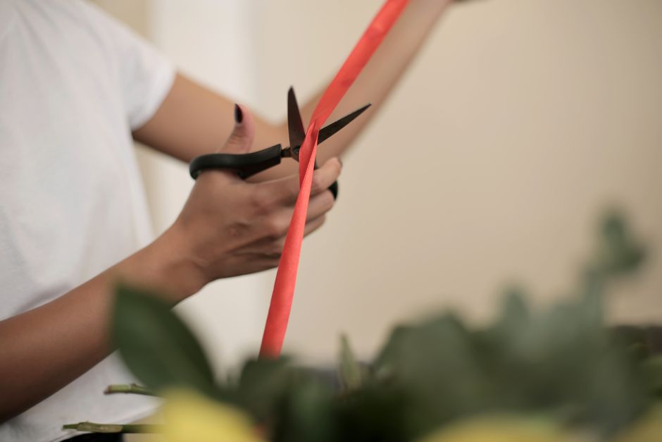 Close-up of a florist's hands holding a pair of black-handled scissors, carefully trimming a coral-colored ribbon on a floral arrangement. The background shows a blurred arrangement of various fresh f