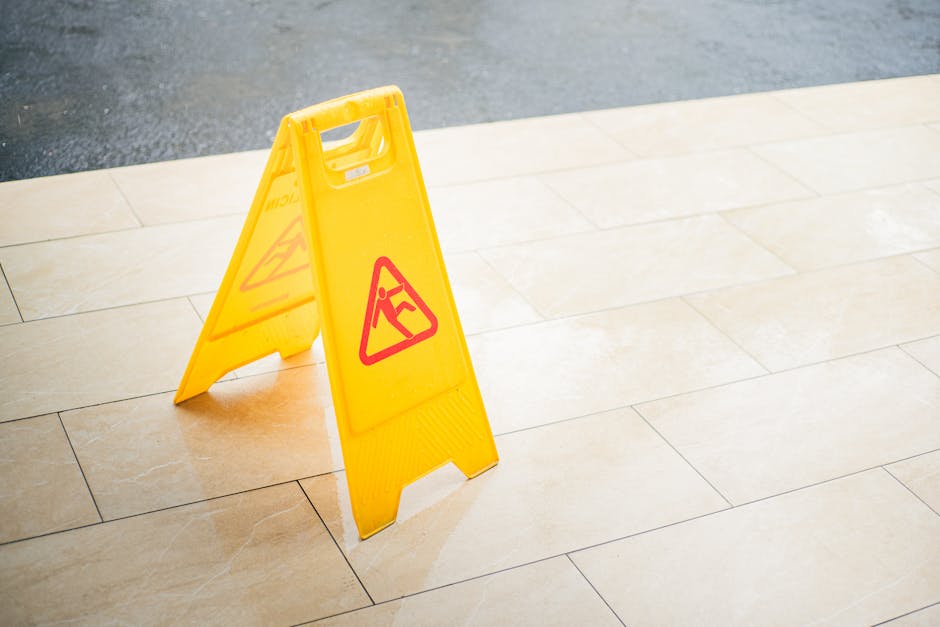 A bright yellow caution wet floor sign with a red warning icon and text, placed on a light beige tiled floor in an indoor environment. The sign is made of plastic and stands upright, indicating a slip