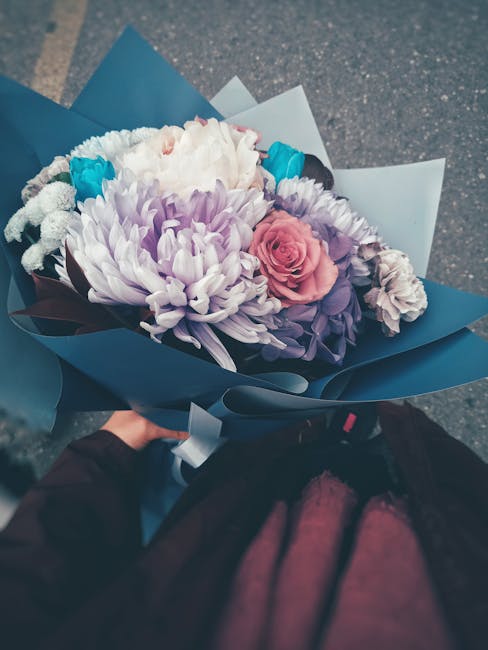 A vibrant floral bouquet held by a person, featuring a variety of fresh flowers including a large white chrysanthemum, soft pink roses, purple lisianthus, and small bright blue blooms, all arranged in