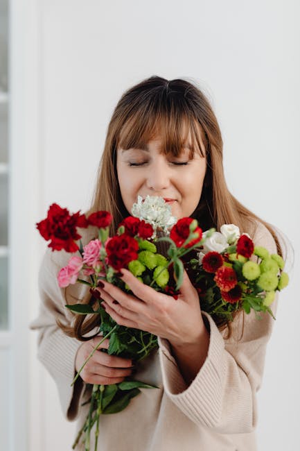 A woman with light brown hair and bangs, wearing a beige sweater, holds a vibrant floral bouquet close to her face with closed eyes and a gentle smile. The bouquet features a mix of fresh flowers, inc