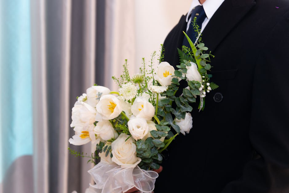A person dressed in a formal black suit and white shirt holding a floral bouquet consisting of white flowers, including roses and lisianthus, accented with green eucalyptus leaves. The bouquet is arra