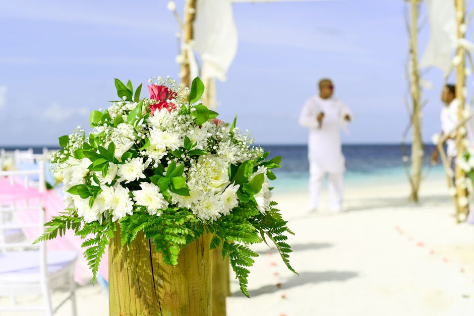A vibrant floral arrangement featuring white roses, lilies, and baby's breath with lush green foliage, arranged in a balanced bouquet. The bouquet is placed in a small wooden container, situated on a