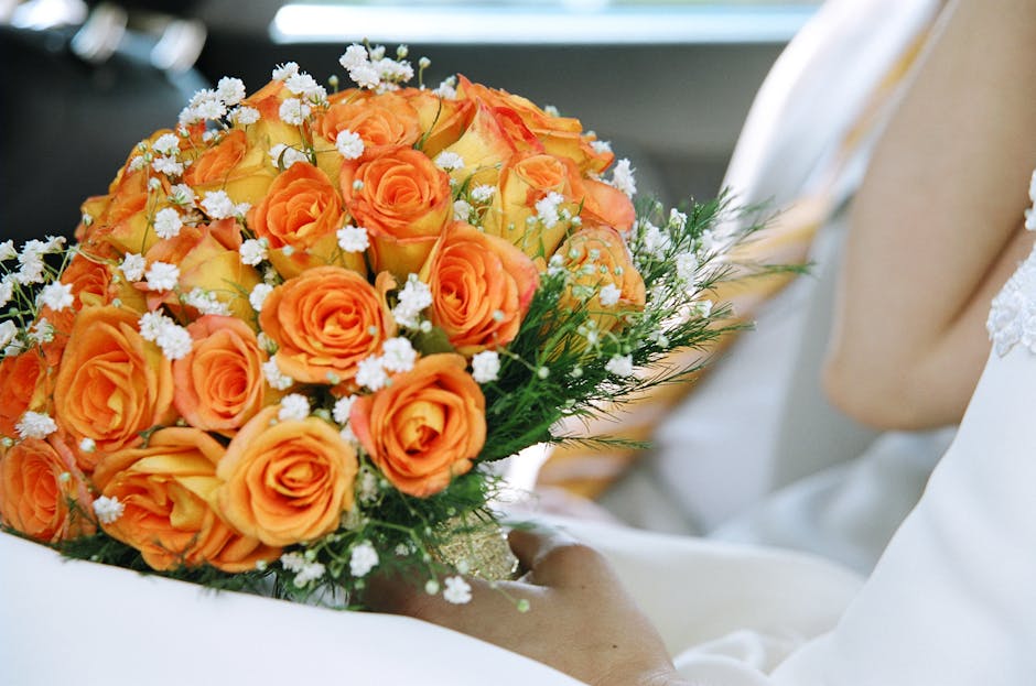 A close-up of a floral bouquet featuring vibrant orange roses with tightly packed petals, complemented by small white filler flowers and lush green foliage. The bouquet is held by a person dressed in
