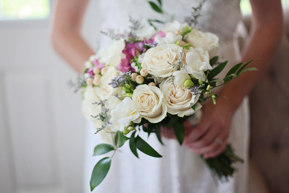 A bride holding a bouquet of fresh floral arrangement featuring large ivory and cream roses, complemented by smaller pink blossoms, white lisianthus, sprigs of lavender, and lush green foliage, wrappe