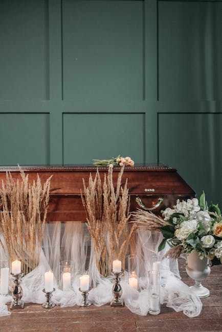 A floral arrangement in front of a wooden coffin with a dark finish, decorated with tall, feathery beige pampas grass and white candles in silver holders, set against a muted green paneled wall. To th