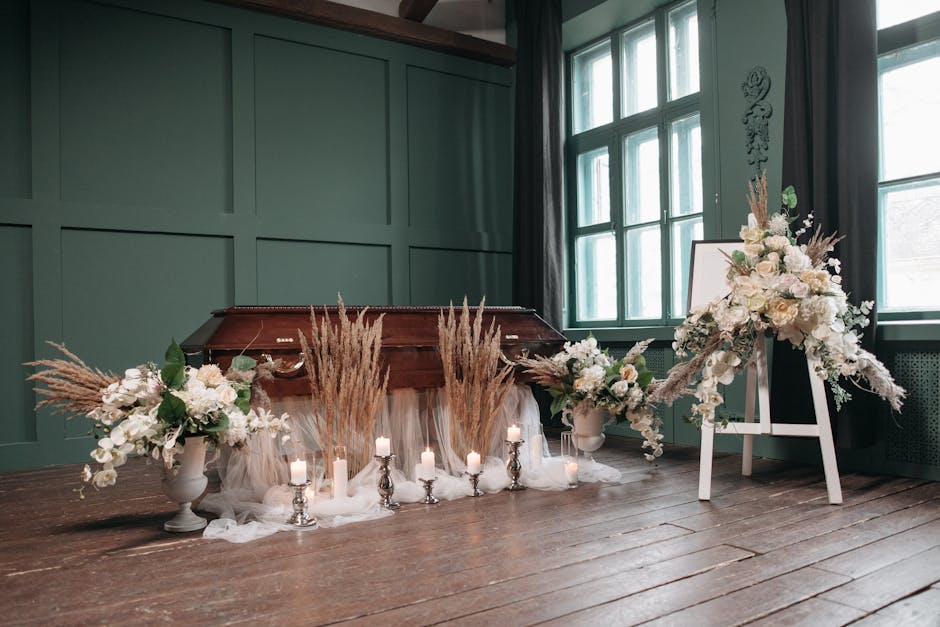 A floral arrangement for a funeral display featuring two large white urn-style vases and a white easel, all adorned with elegant white and cream flowers including roses, lilies, and hydrangeas. The ar