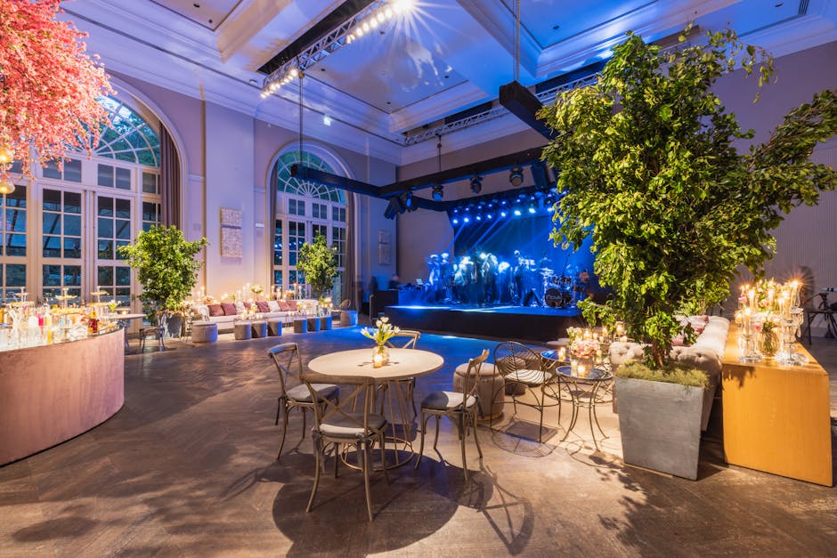 Inside a spacious, elegantly decorated event venue featuring large arched windows and high ceilings illuminated with soft, warm lighting. The room is arranged with round tables topped with small flora