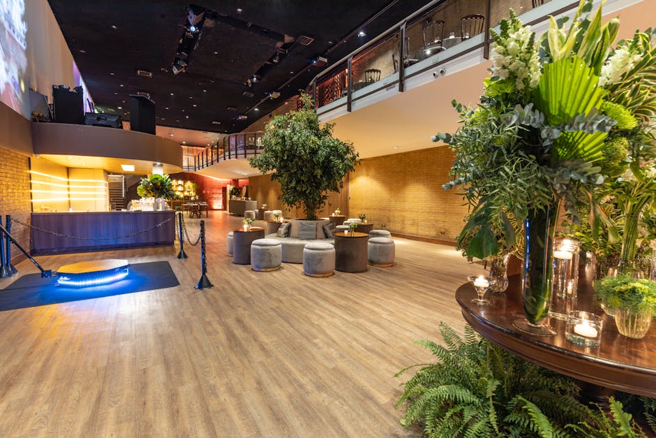 Inside the Queens Theatre Hornchurch event space, a floral arrangement featuring white lilies, lush green foliage, and tall stems is displayed in a glass vase on a polished wooden table. The arrangeme