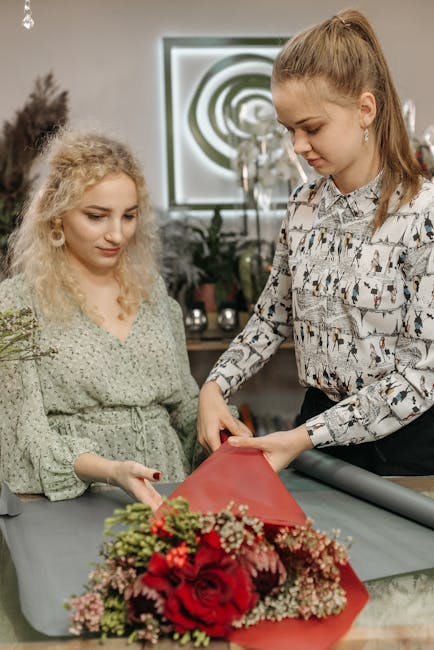 Two women are engaged in floral arrangement, with one woman holding a bouquet wrapped in red tissue paper and the other observing attentively. The bouquet features large red roses, complemented by sma